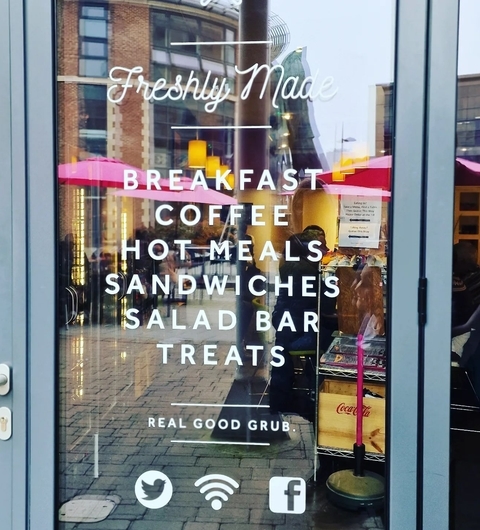 Glass entrance of Cafe YumYum featuring a menu of offerings: breakfast, coffee, hot meals, sandwiches, salad bar, and treats.