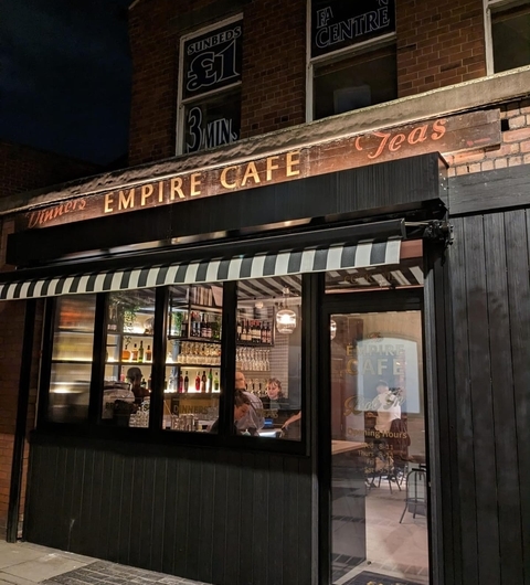 Cozy exterior of The Empire Cafe at night, featuring a striped awning and warm interior lights illuminating the bar.