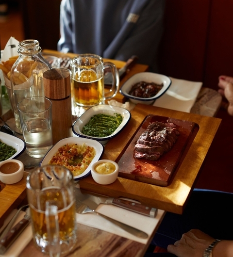 A wooden table set with grilled steak, sauces, and sides, alongside glasses of beer and water, creating a cozy dining atmosphere.