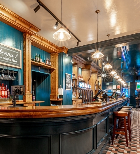 Warm wooden bar with polished counter, illuminated by pendant lights, surrounded by shelves of bottles and a checkered floor.