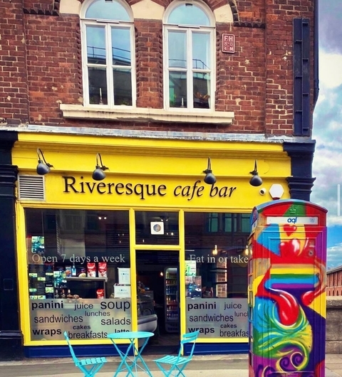 Bright yellow café exterior with large windows, colorful signage, and vibrant blue chairs outside, set against a brick wall.