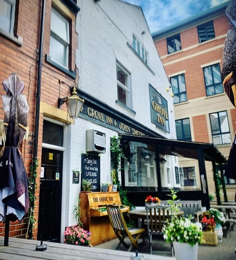Charming pub exterior with wooden seating, flower pots, and a welcoming sign, set against a bright blue sky.