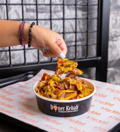 A hand lifts a portion of loaded doner kebab from a bowl, surrounded by fresh toppings, on a branded tray.