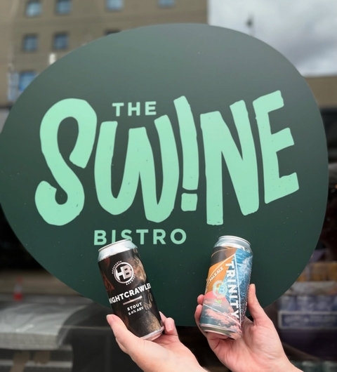 Two hands hold craft beer cans in front of The Swine Bistro's vibrant green sign, showcasing a lively dining atmosphere.