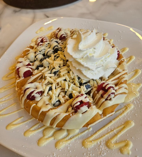 A waffle topped with fresh raspberries, blueberries, white chocolate shavings, white chocolate sauce drizzle, icing sugar and a swirl of whipped cream at 39 Desserts, Leeds.
