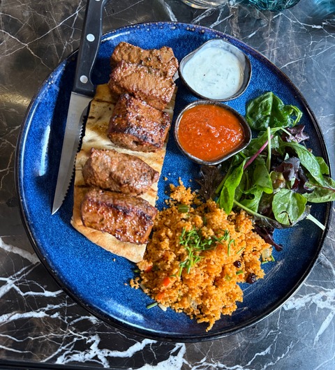 A grilled lamb skewer on flatbread with cous cous, tomato sauce, herb yogurt and a side salad on a deep blue plate at Izgara Mediterranean Grill, Garforth, Leeds.