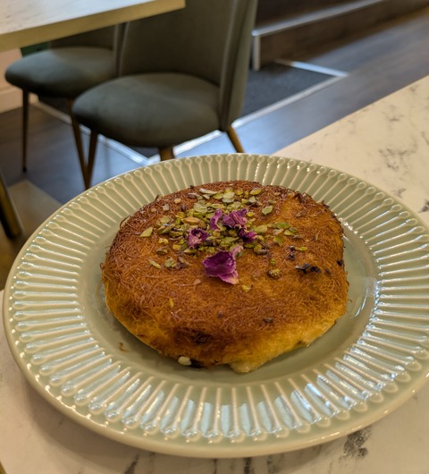 A round kunafa pastry topped with chopped pistachios and rose petals, served on a green plate at Jasmine Cafe, Leeds.