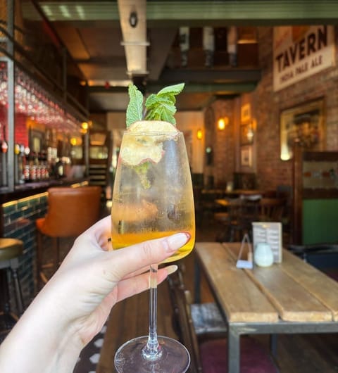 Hand holding a spritz cocktail with mint garnish in front of The Clock Inn bar interior with warm lighting and vintage TAVERN sign
