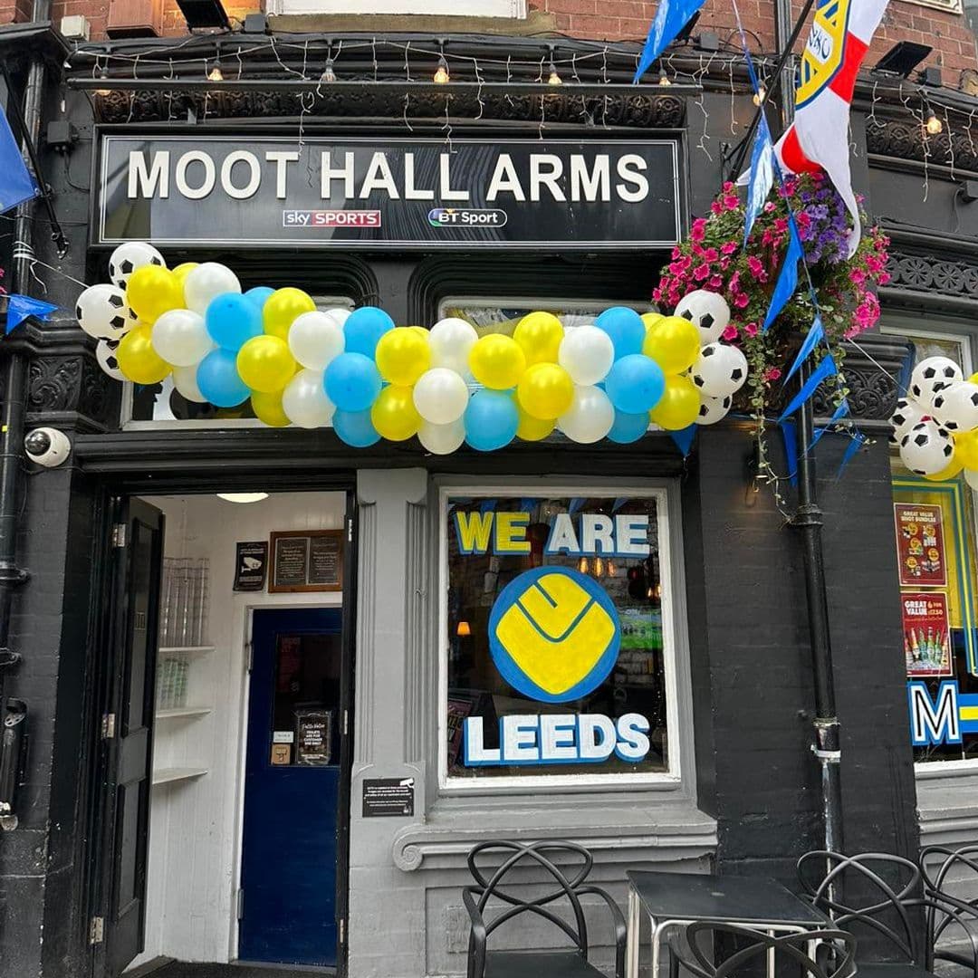 Alt text: Entrance of The Moot Hall Arms pub decorated with blue, yellow, and white balloons, featuring a sign that reads "WE ARE LEEDS" in the window. The exterior has flags and flowers, creating a welcoming atmosphere.