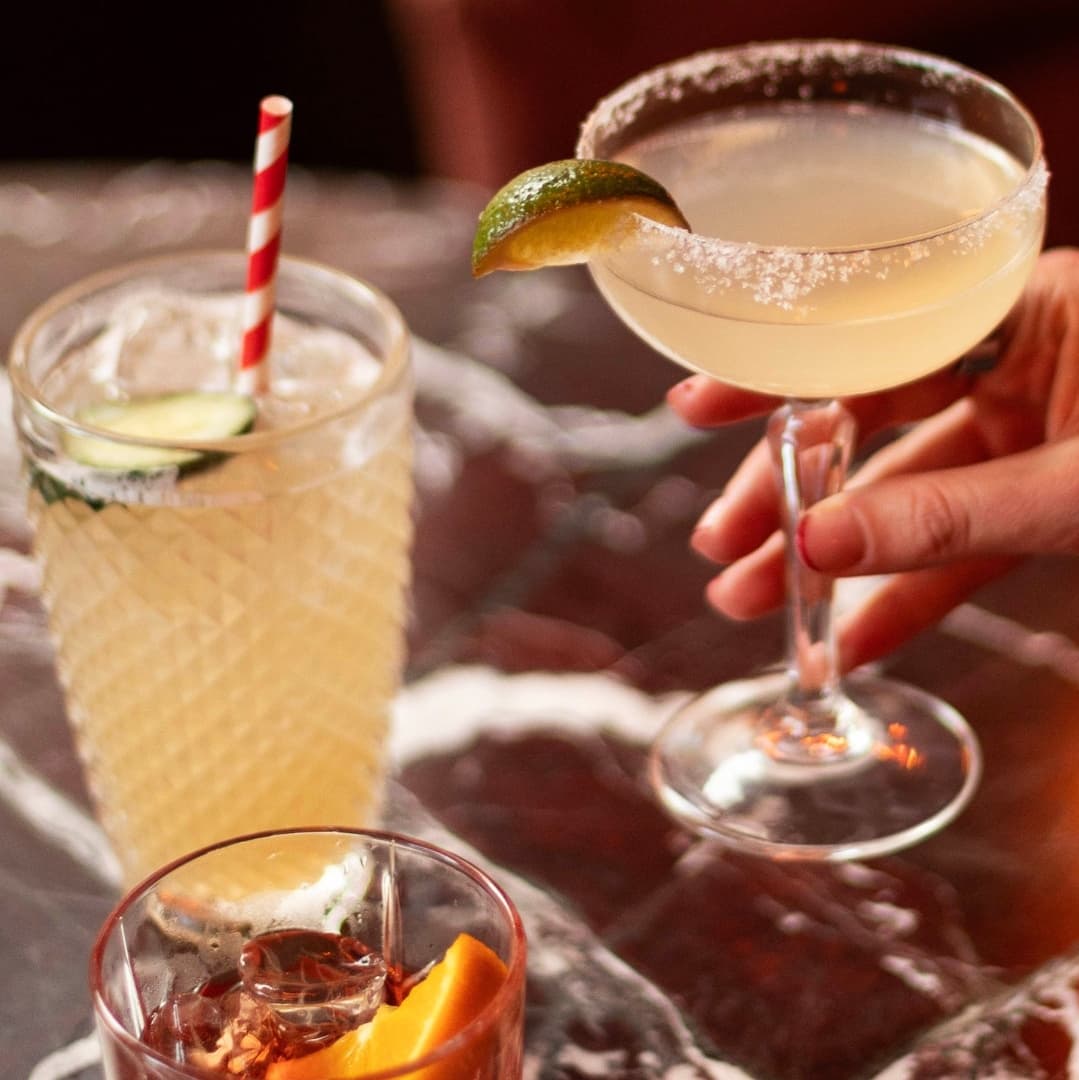 A hand holds a salted margarita with a lime wedge, alongside two other colourful cocktails on a marble table.