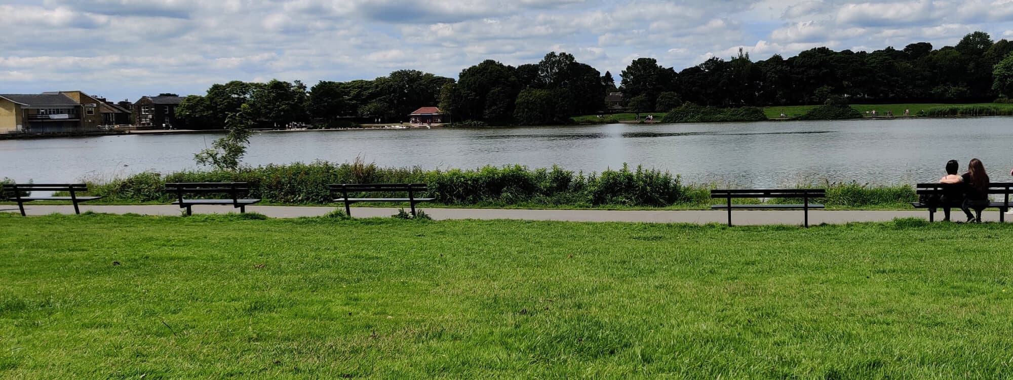 Green grass and lake in the background with trees behind. Couple sat on a bench to the right.