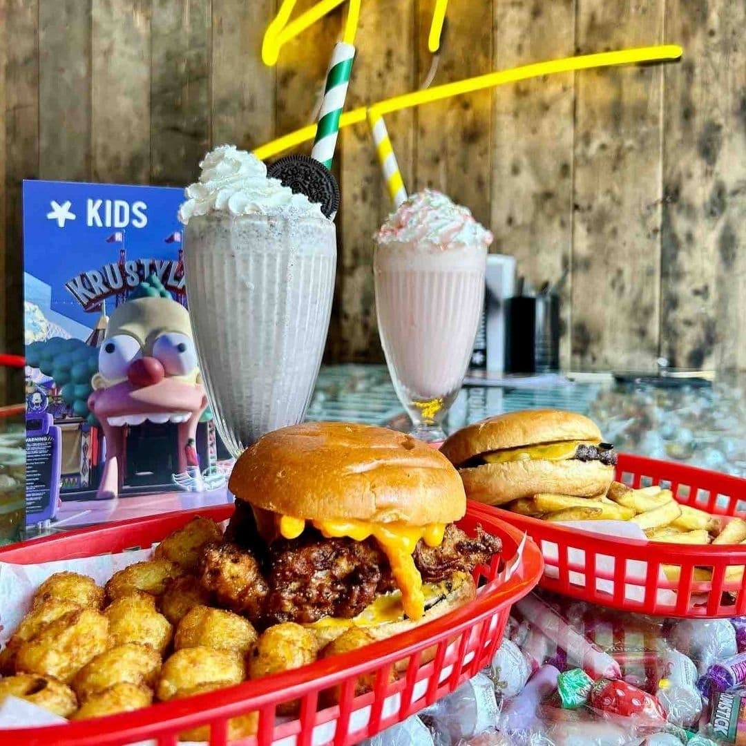 Delicious dining experience at Almost Famous, featuring a juicy cheeseburger with tater tots, a classic hamburger with fries, and two creamy milkshakes topped with whipped cream, one garnished with an Oreo cookie. A kids' menu card with a cartoon character is visible in the background, adding a fun, family-friendly vibe.