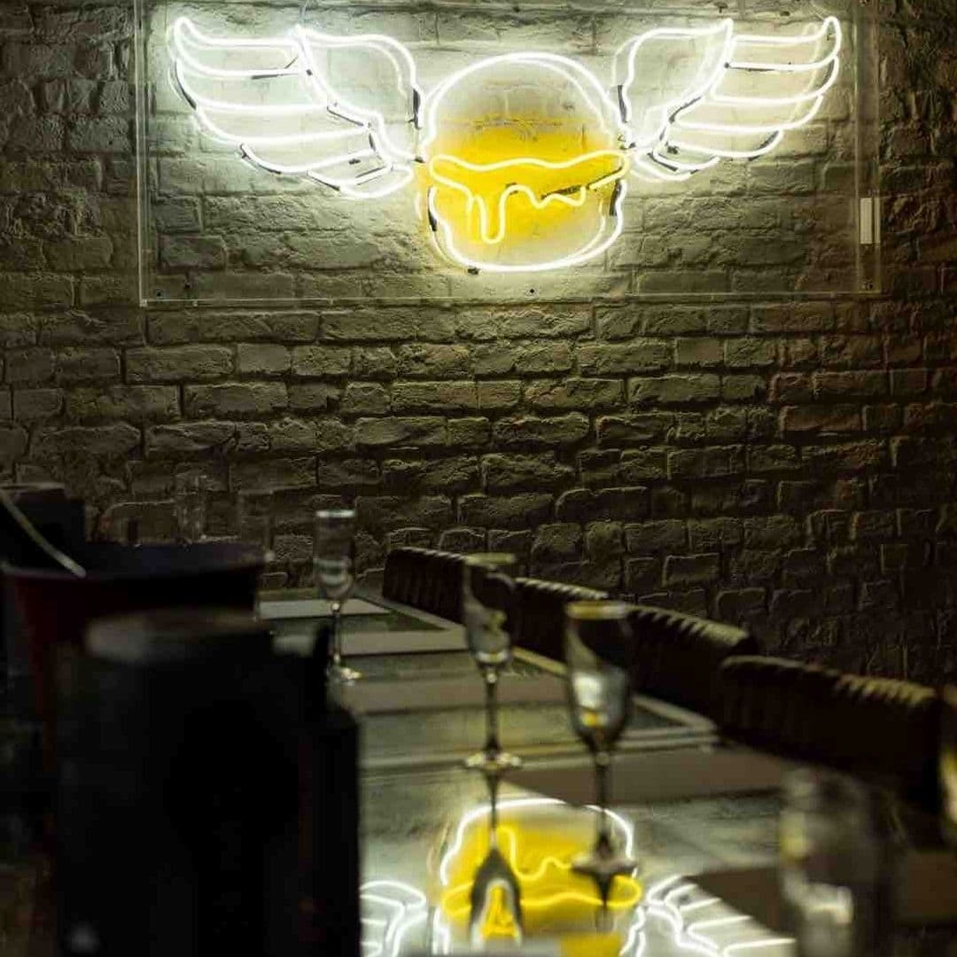 Neon winged egg sign on brick wall at Almost Famous with reflections on a glass-topped bar counter.
