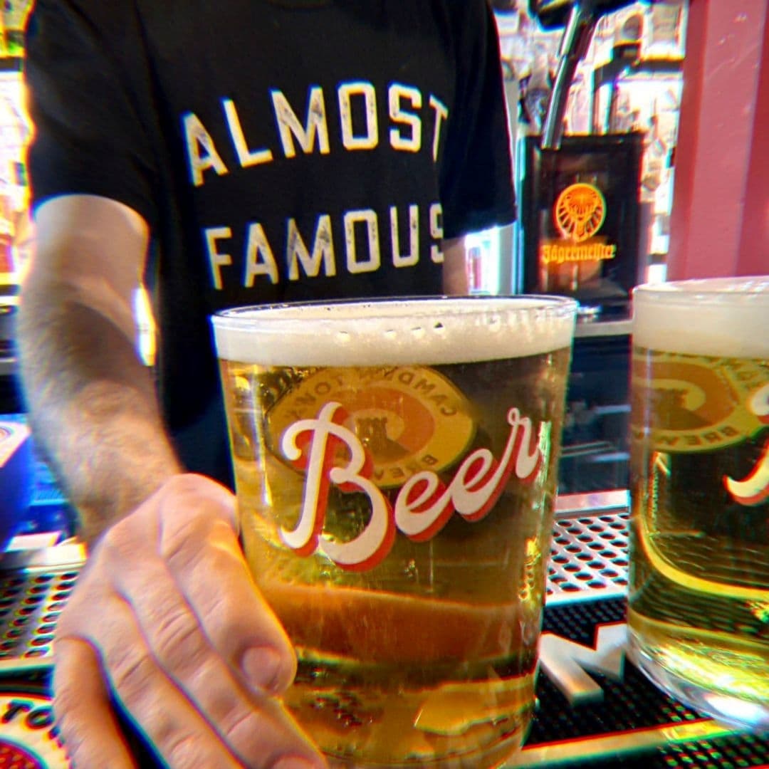 Person serving a beer at Almost Famous with a bartender wearing a branded t-shirt in the background.