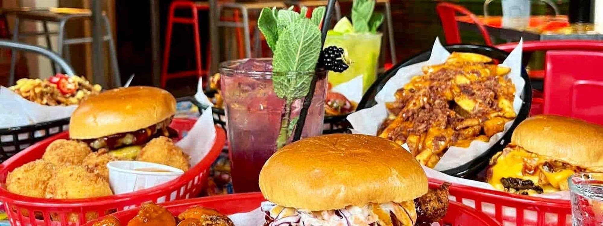 Mouthwatering array of American diner food at Almost Famous, featuring juicy burgers in buns, crispy fried chicken, loaded cheese fries, and refreshing cocktails garnished with fresh mint.