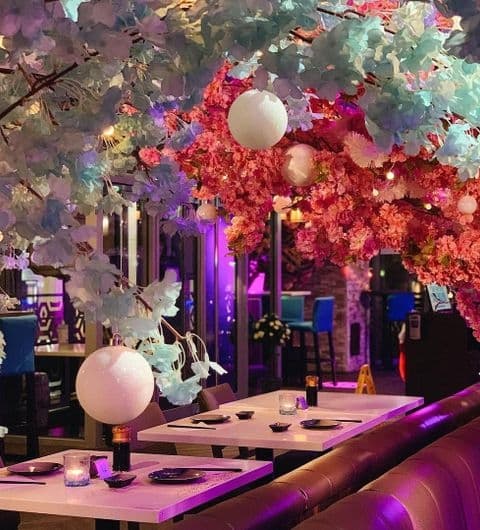 Interior of Blue Sakura featuring an elegant dining area with tables set for guests under a canopy of vibrant pink and blue artificial cherry blossoms, with spherical white lanterns hanging amidst the foliage, creating a cozy and enchanting atmosphere.