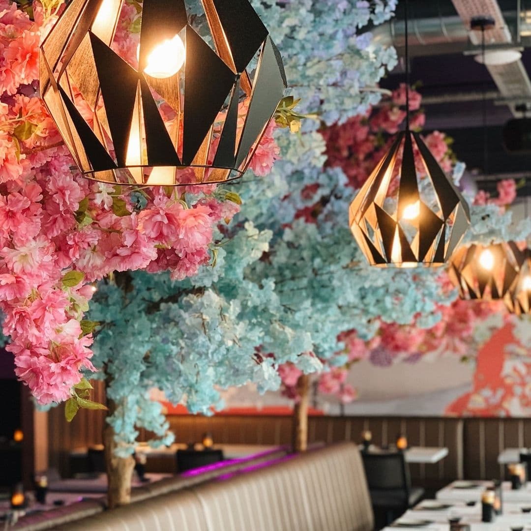 Elegant interior of Blue Sakura featuring geometric pendant lights and vibrant pink and blue artificial cherry blossoms over a cozy dining area with modern furnishings.