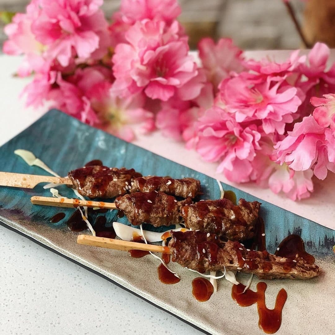 Grilled skewered meat with a glaze, sprinkled with sesame seeds, served on a blue ceramic plate at Blue Sakura, with vibrant pink cherry blossoms in the background.