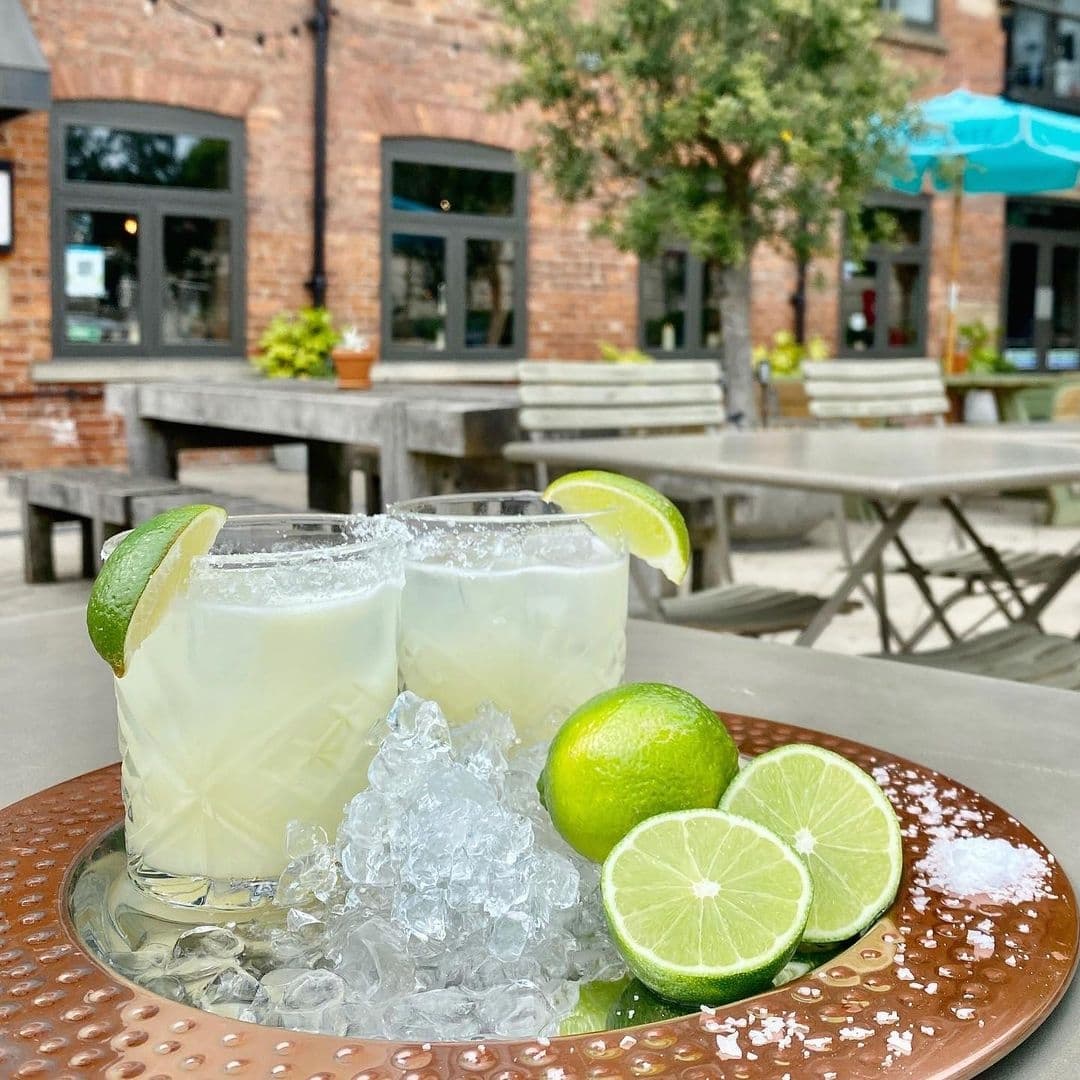 Two refreshing margarita cocktails with lime wedges on a copper tray, crushed ice scattered around, with outdoor seating of Brasserie Blanc in the background.