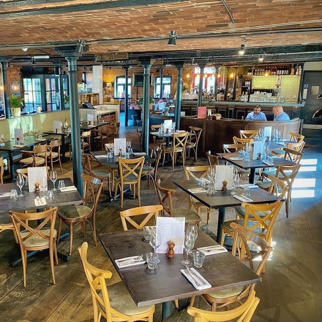 Interior of Brasserie Blanc with wooden tables set for dining, exposed brick ceiling, and patrons seated at the bar area.