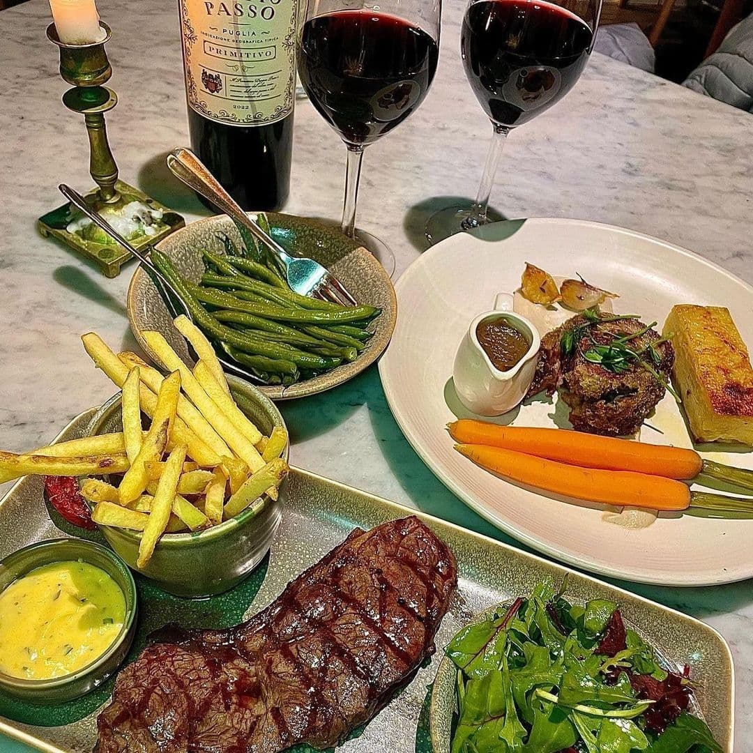 Dining experience at Brasserie Blanc featuring a bottle of red wine, two filled glasses, and an array of dishes including a grilled steak with fries and béarnaise sauce, green beans, a plate with roasted vegetables and meat, and a fresh salad.