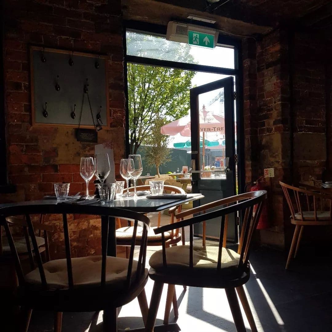 Interior of Brasserie Blanc with sunlight streaming through an open door, highlighting empty wine glasses set on a table, with exposed brick walls and a view of outdoor seating.