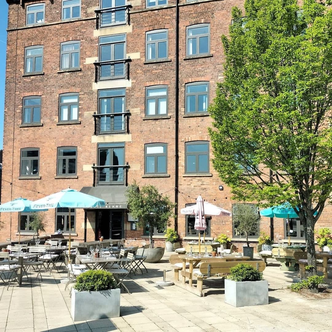 Outdoor seating area at Brasserie Blanc with wooden tables and chairs, sun umbrellas, and green trees on a sunny day, set against a brick building façade.