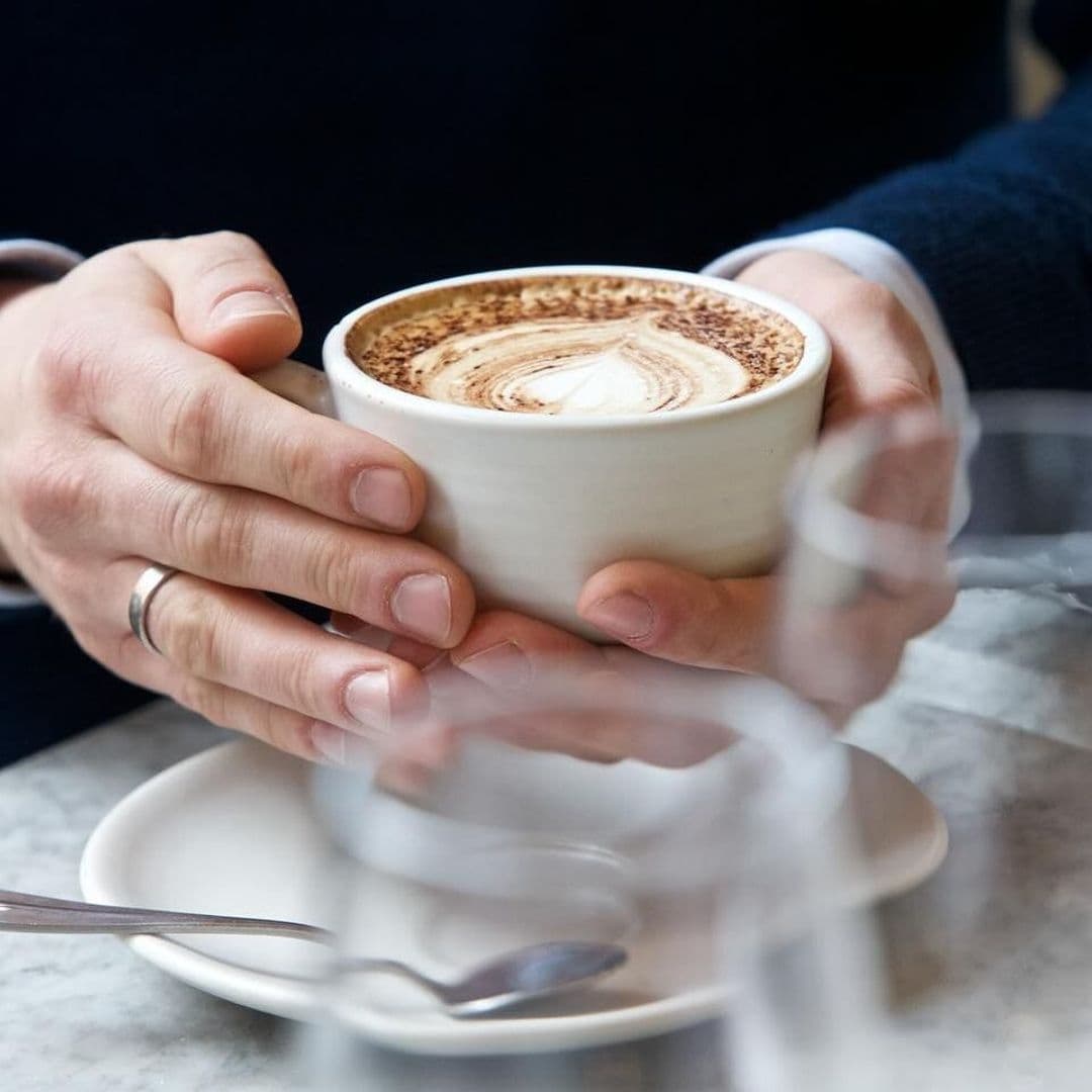 Close-up of a person's hands holding a cup of cappuccino with artful foam design at Brasserie Blanc.