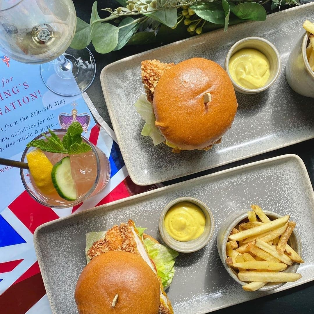 Alt text: "Delicious meal at Brasserie Blanc featuring two gourmet burgers with lettuce on brioche buns, a side of golden fries, a bowl of mustard, and a refreshing cocktail garnished with a mint leaf and citrus slices."