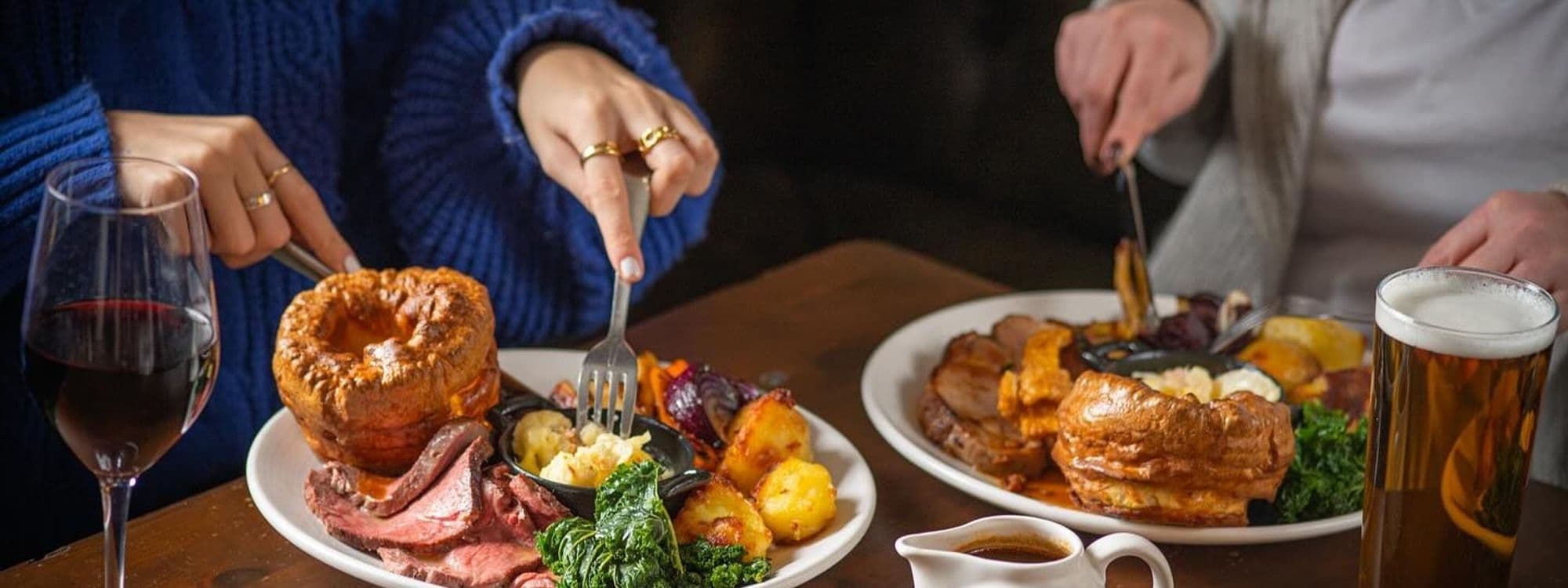 Alt text: "Diners enjoying a hearty meal at Brasserie Blanc with a glass of red wine and a pint of beer, featuring roast beef, Yorkshire puddings, roasted potatoes, and green vegetables."