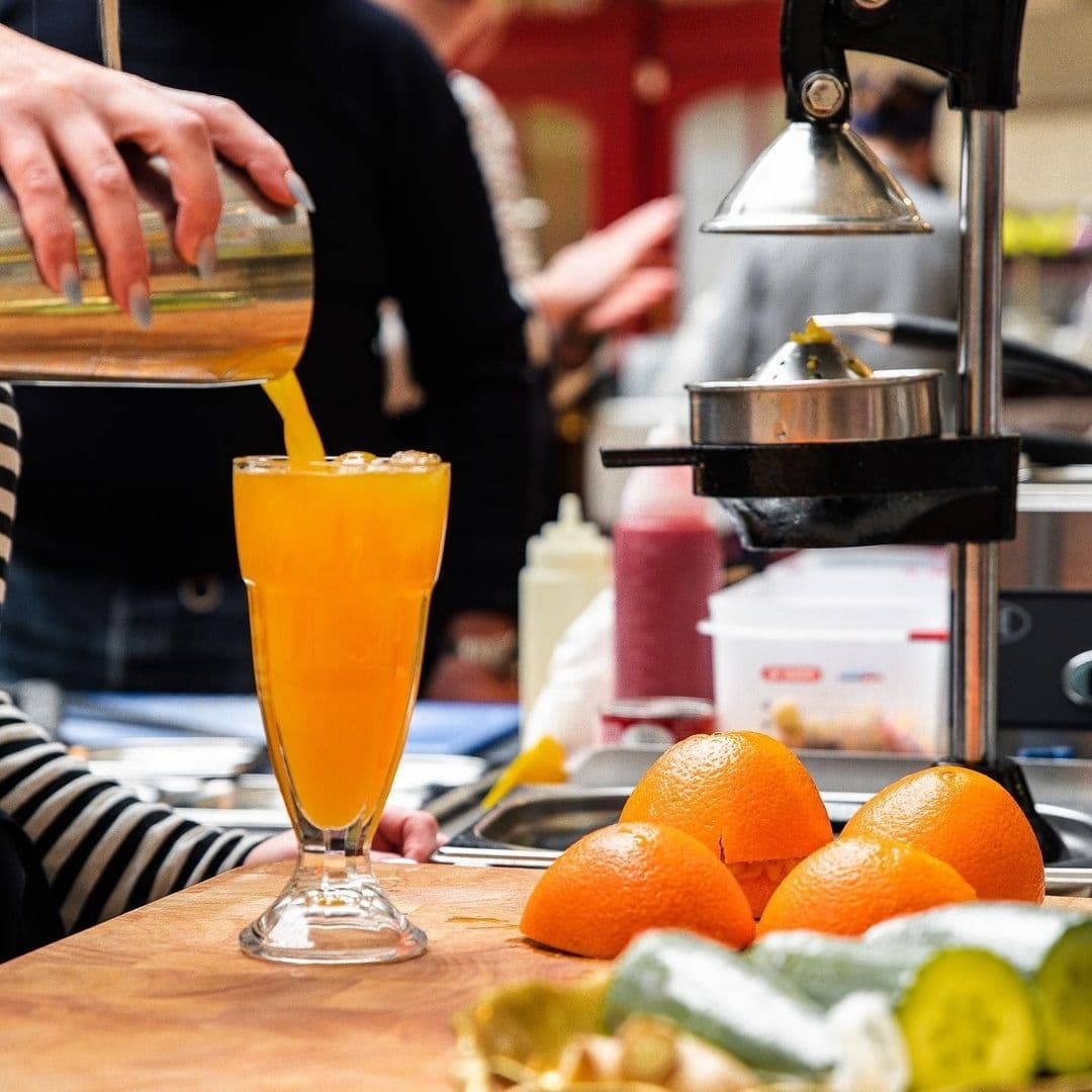 Freshly-squeezed orange juice being poured into a glass at Bruschetta, with whole oranges and a citrus juicer in the background.