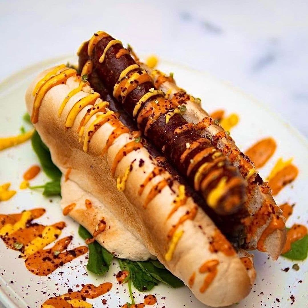 Gourmet hot dog with condiments on a bed of greens, served on a white plate at Bruschetta.