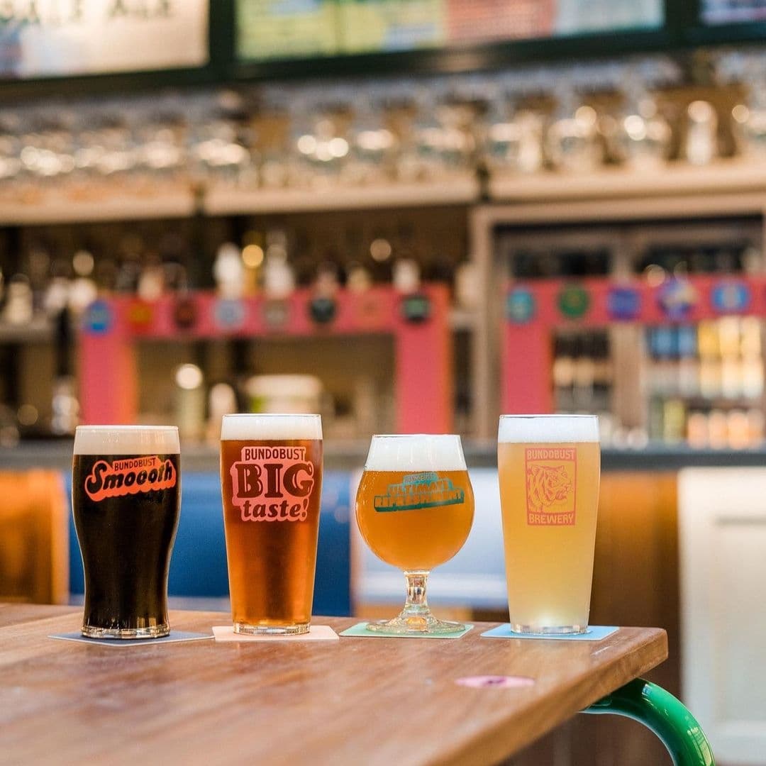Alt text: "Four different styles of craft beer in branded glasses on a wooden bar at Bundobust, with a blurred view of the bar area in the background."