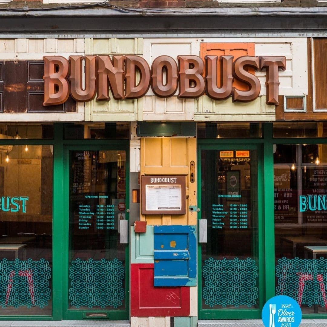 Exterior view of Bundobust restaurant with large 3D letter signage above the entrance, green framed windows and doors, and a menu board next to a multi-colored post box.