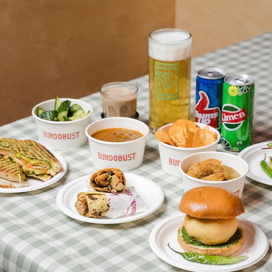Assorted Indian street food and beverages on a checkered tablecloth at Bundobust, featuring sandwiches, a burger, samosas, snacks in bowls, a glass of beer, and canned drinks.