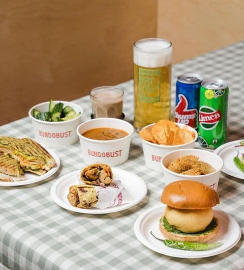 Assorted Indian street food and drinks on a checkered tablecloth at Bundobust, including a sandwich, a burger, bowls of curry, snacks, a pint of beer, and canned beverages.
