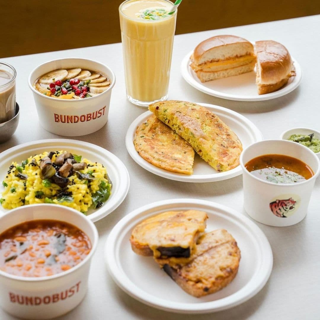 Assorted Indian dishes and a drink on a table at Bundobust, featuring bowls of curry and rice, stuffed bread, a sandwich, and a tall glass of a yellow beverage.