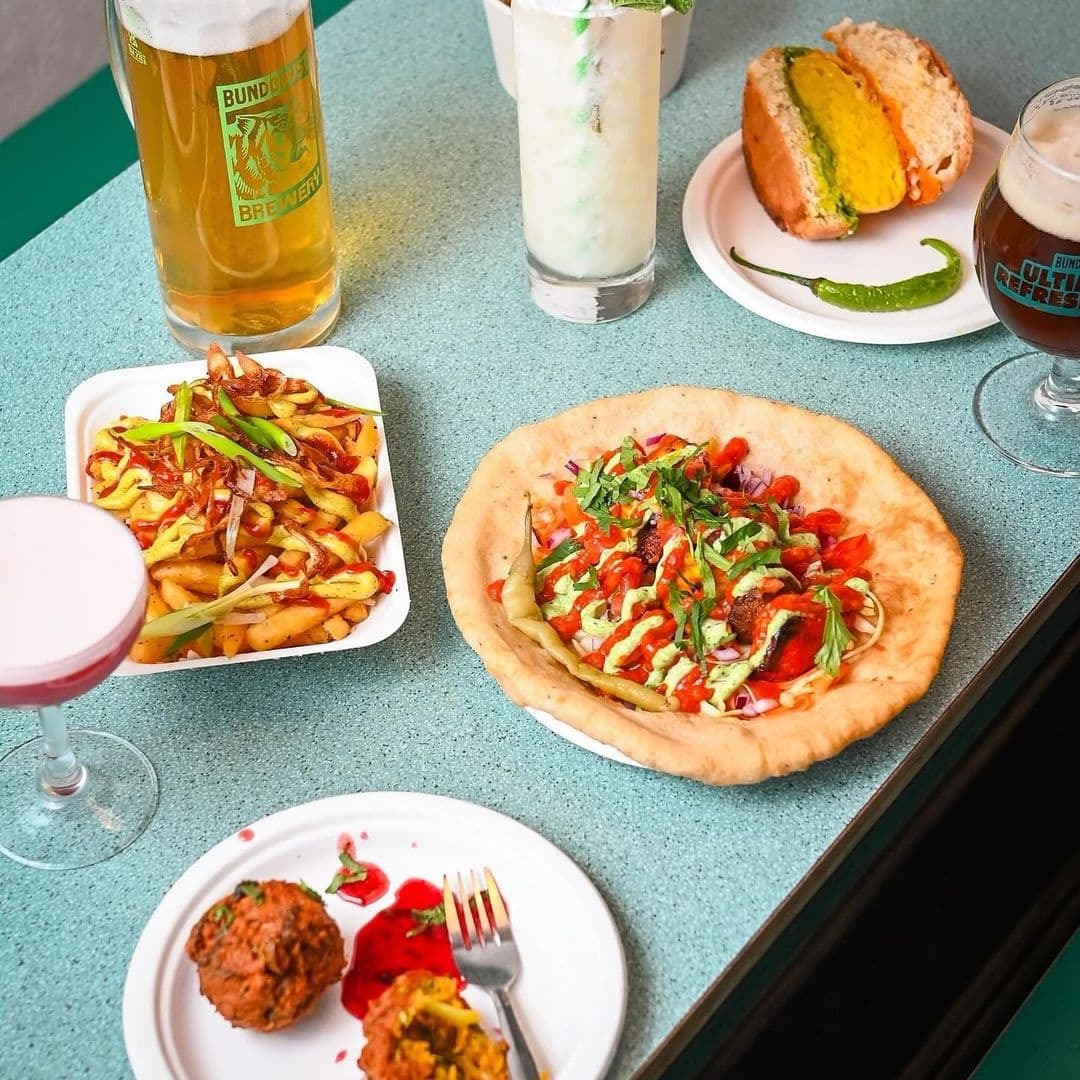 Assorted Indian street food dishes and drinks on a table at Bundobust, featuring a pint of beer, a cocktail, masala fries, a stuffed bread roll, and a flatbread topped with colorful toppings.