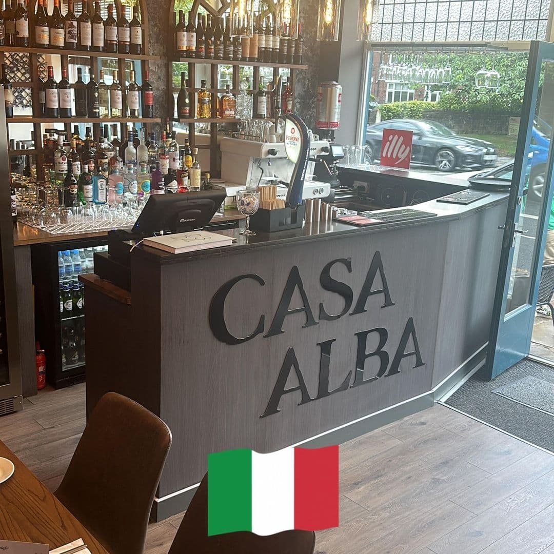 Interior view of Casa Alba featuring a well-stocked bar with various bottles, an espresso machine with the Illy logo, and the establishment's name prominently displayed on the front counter. An Italian flag is visible in the foreground.