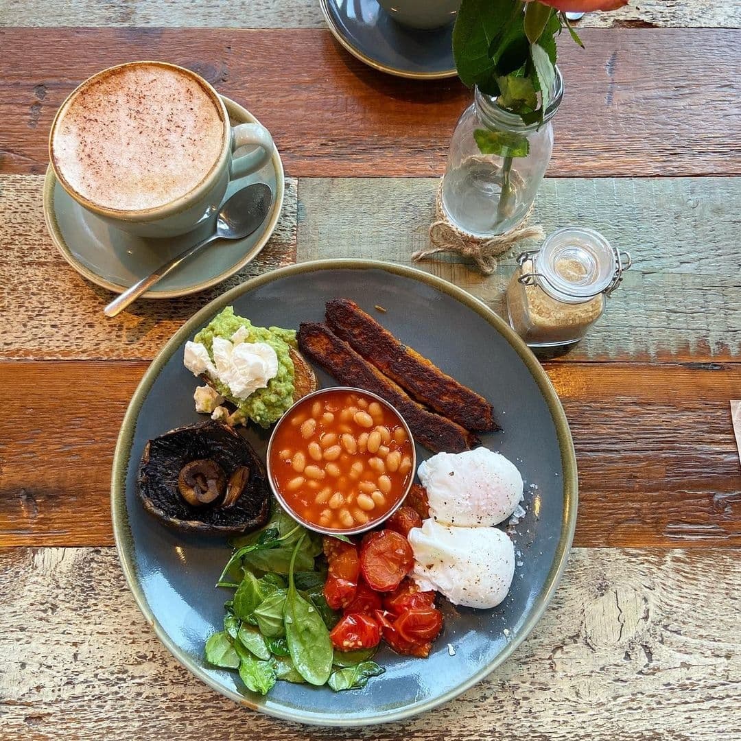 Alt text: "Delicious breakfast served at Cha Lounge featuring poached eggs, baked beans, roasted tomatoes, spinach, avocado, mushrooms, bacon strips, and a cup of cappuccino on a rustic wooden table."