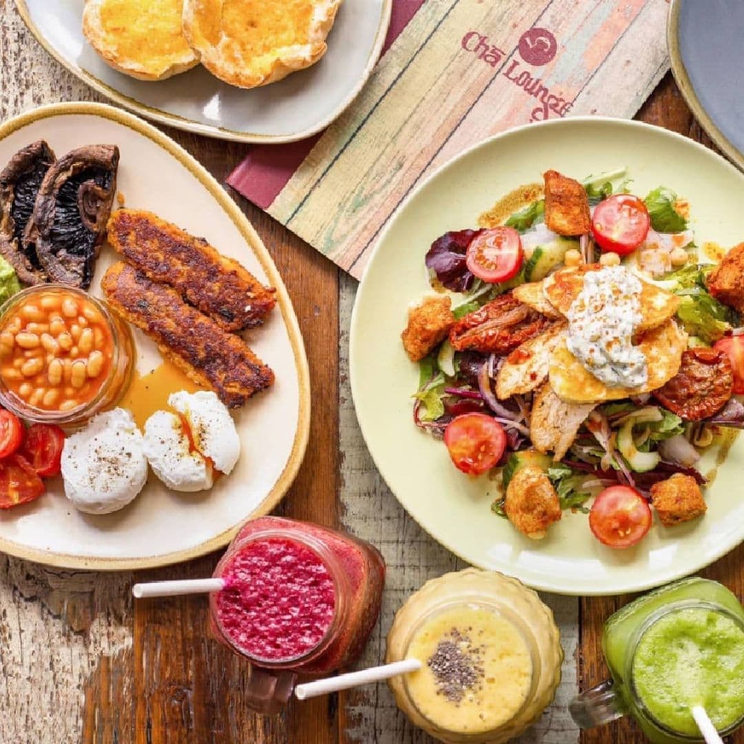 Assorted breakfast dishes and smoothies on a wooden table at Cha Lounge, featuring poached eggs, beans, grilled plantains, salad with crispy croutons, and freshly baked bread.