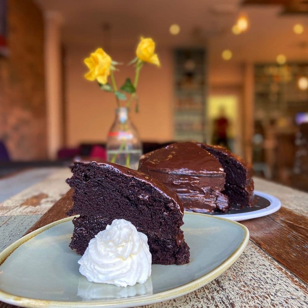 A slice of rich chocolate cake with a dollop of whipped cream on a ceramic plate, with a full cake in the background, at Cha Lounge. Yellow roses in a vase add a vibrant touch to the cozy ambiance.