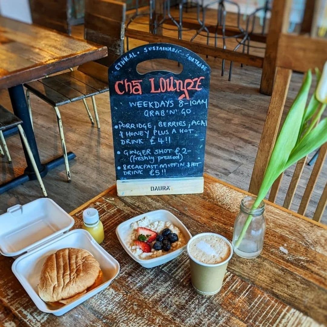 Alt text: "Cozy breakfast setup at Cha Lounge featuring a chalkboard menu with healthy options, a sandwich in a takeout box, a bowl of porridge with berries, a cup of coffee, and a small bottle of juice on a rustic wooden table."