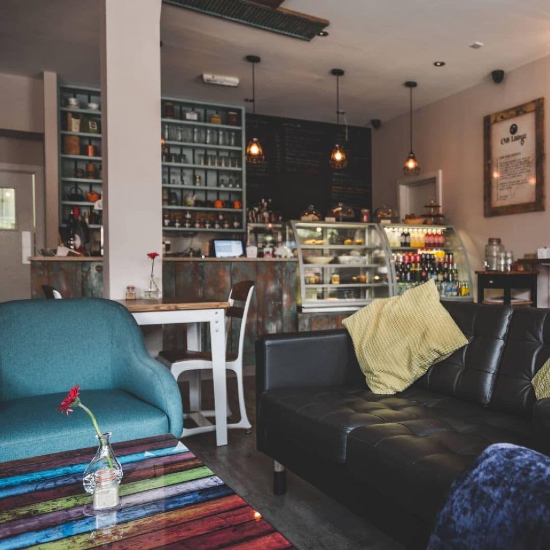 Interior of Cha Lounge featuring comfortable seating with a blue sofa and black leather couch, a colorful striped rug, a rustic bar counter with shelves of bottles and glasses, and a menu board in the background.