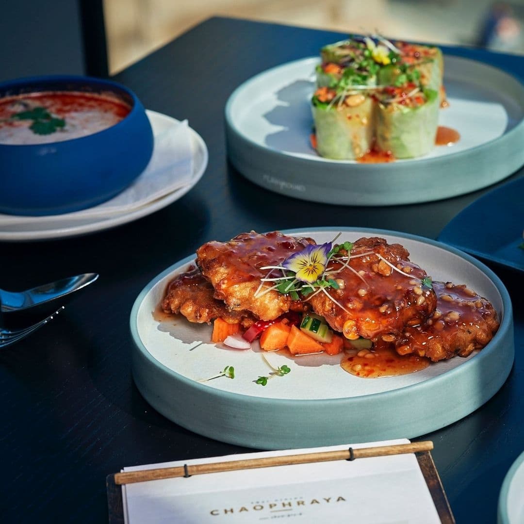 Gourmet dishes on a table at Chaophraya, featuring a plate of crispy chicken with sauce and garnished with a flower, alongside a fresh spring roll and a bowl of soup.