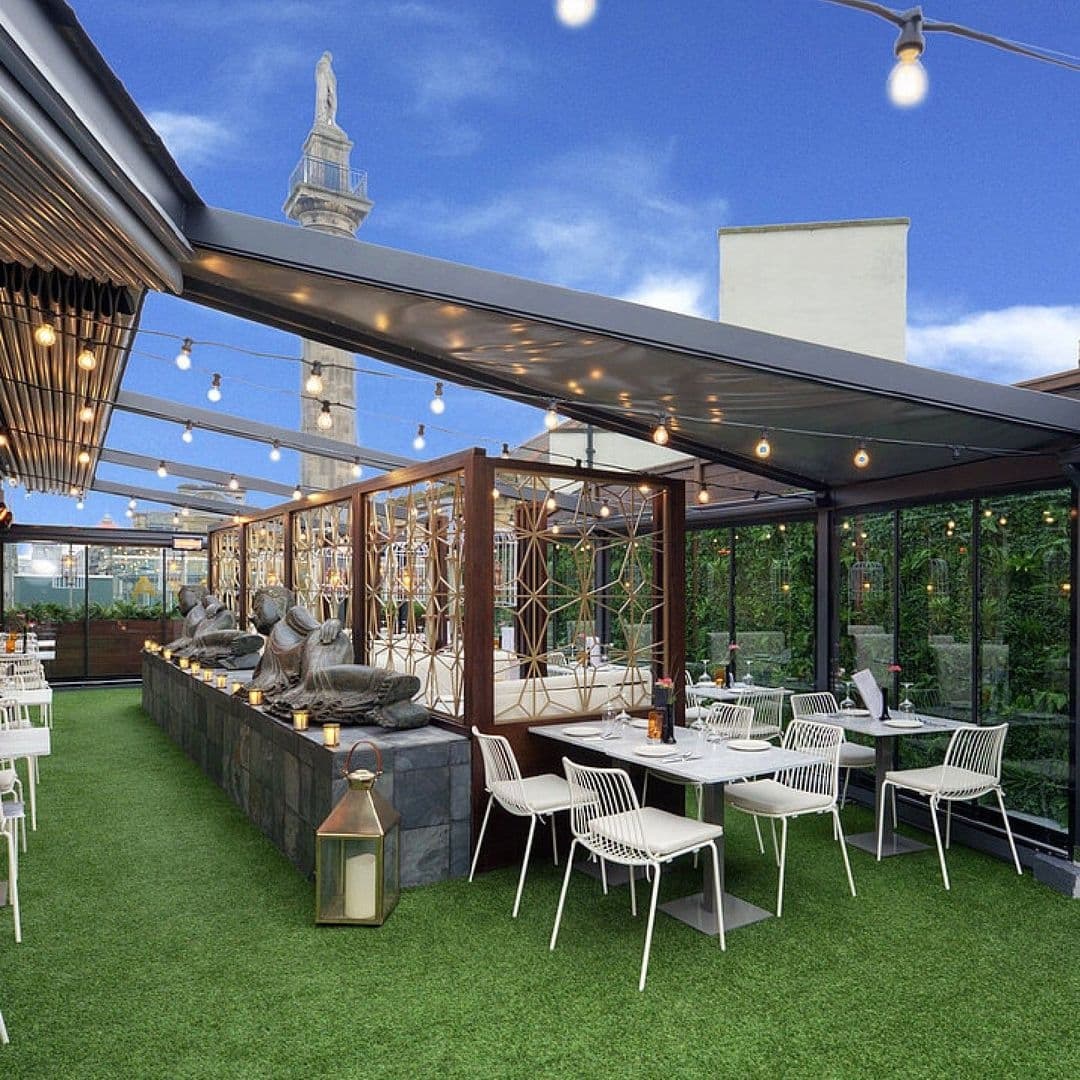 Outdoor rooftop dining area at Chaophraya with white chairs, green artificial grass, string lights, and a view of a statue.