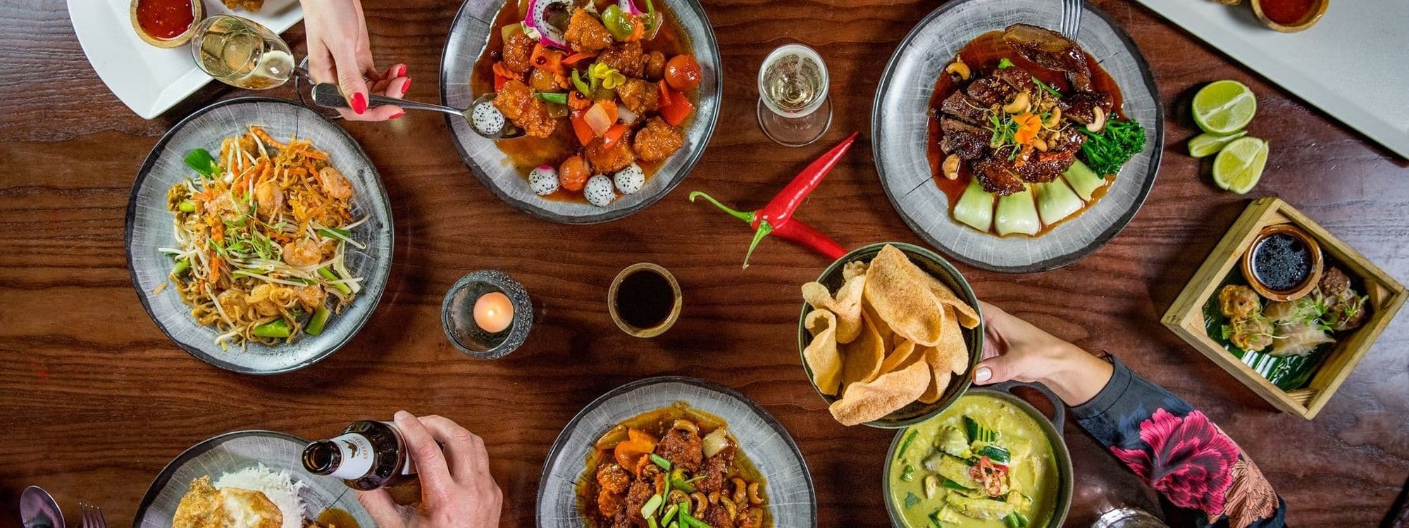 Assorted Thai dishes spread on a wooden table at Chaophraya, featuring pad thai, crispy pork belly, spicy chicken, shrimp dumplings, and green curry, with diners' hands visible, sharing a communal meal.