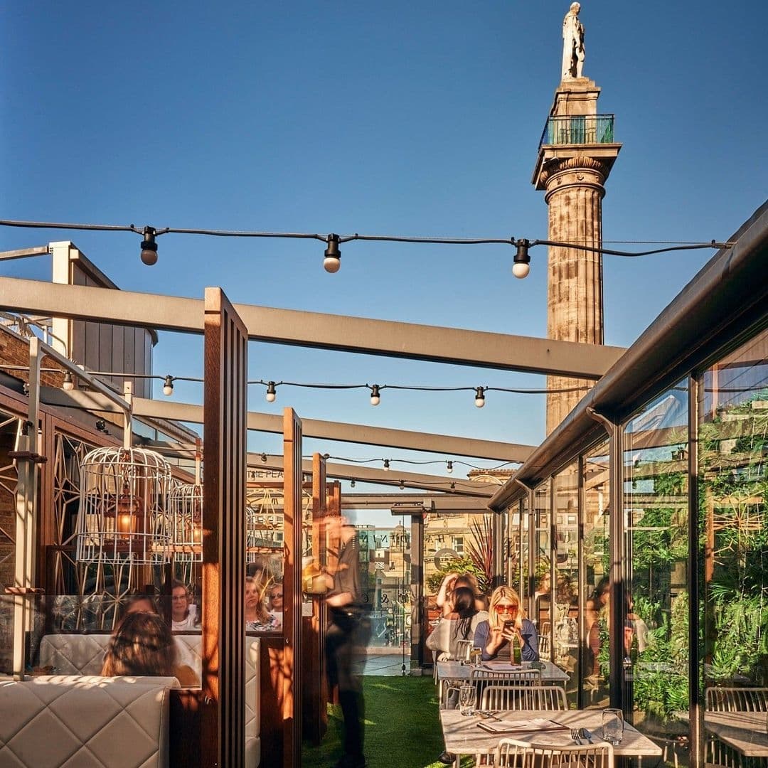 Outdoor rooftop dining area at Chaophraya with guests enjoying meals, string lights above, and a historic column in the background under a clear blue sky.