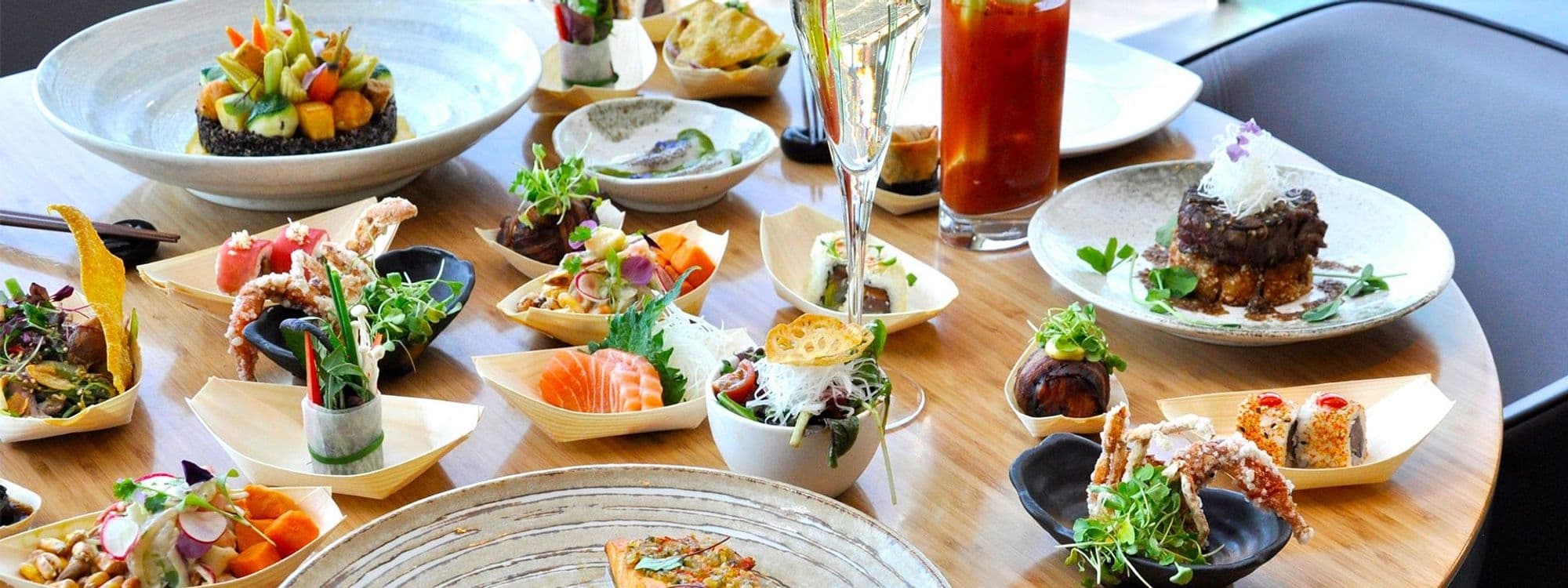 Assorted gourmet dishes on a wooden table at Chino Latino, featuring sushi, ceviche, and various appetizers with vibrant garnishes, accompanied by a glass of champagne and a Bloody Mary cocktail.