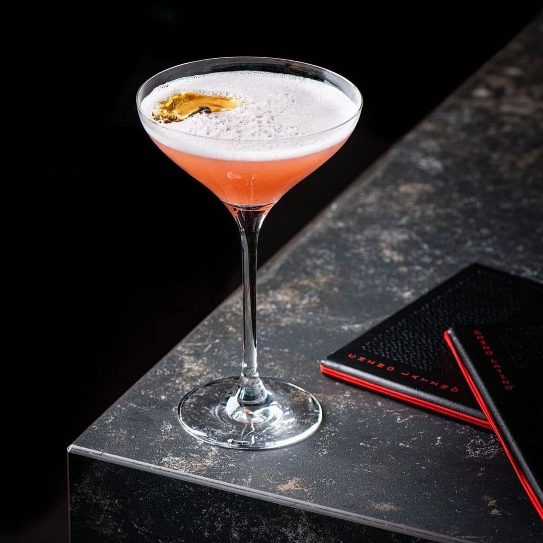 Elegant pink cocktail in a martini glass garnished with an orange peel, presented on a dark bar counter with a menu titled "Chino Latino" next to it.